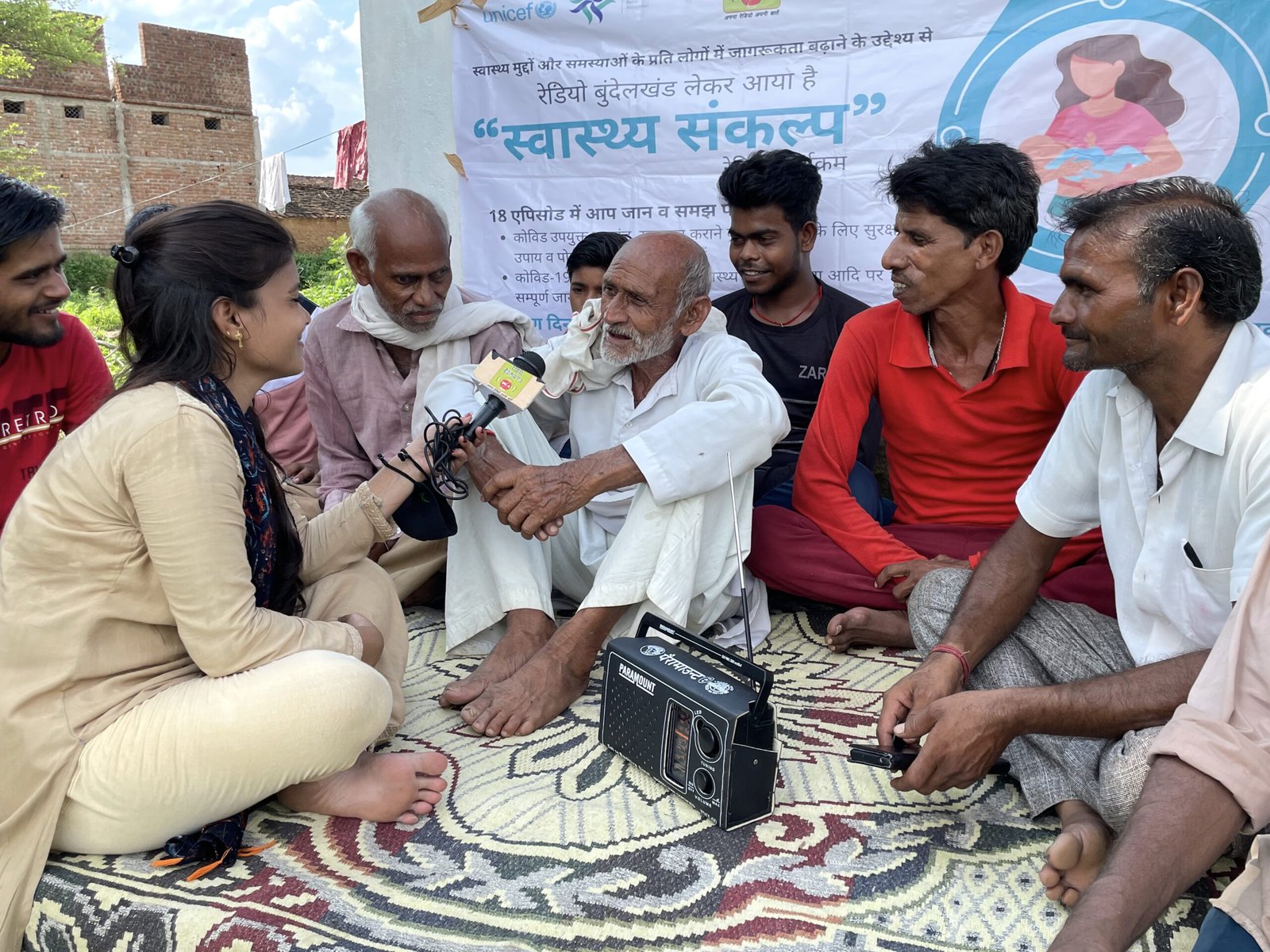 A young reporter talks to the men in the community during a public health camp conducted by Bundelkhand Radio. They are sitting together outdoors on a rug.