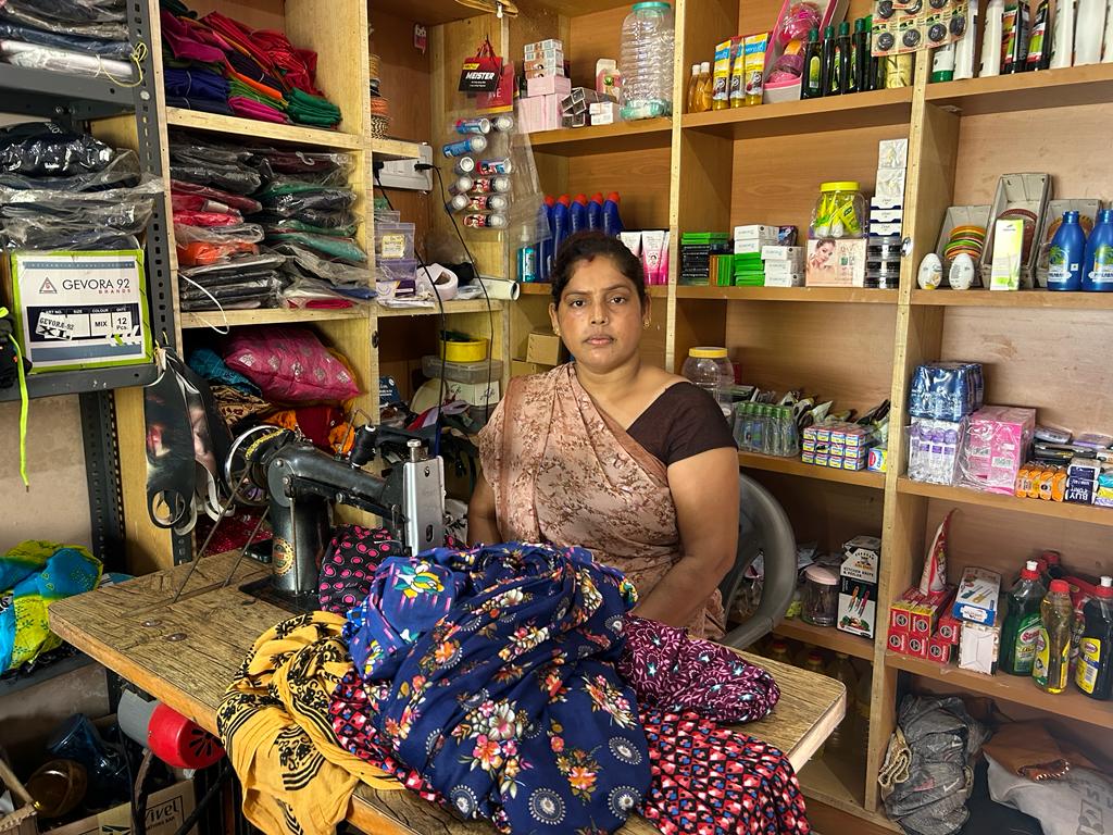 A woman sits in her tailor shop with a sewing machine and fabric in front of her.