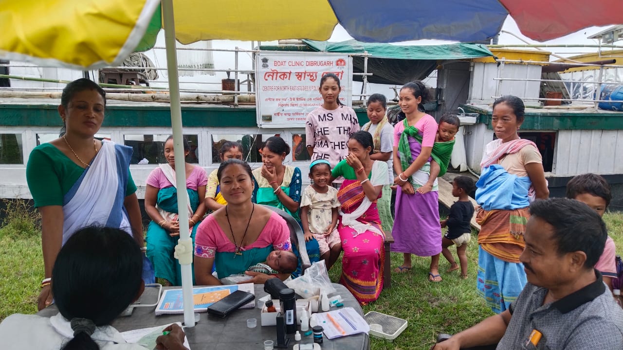 Boat clinic staff conducts an awareness program on family planning. A group of people are huddled under an umbrella with the boat behind them.