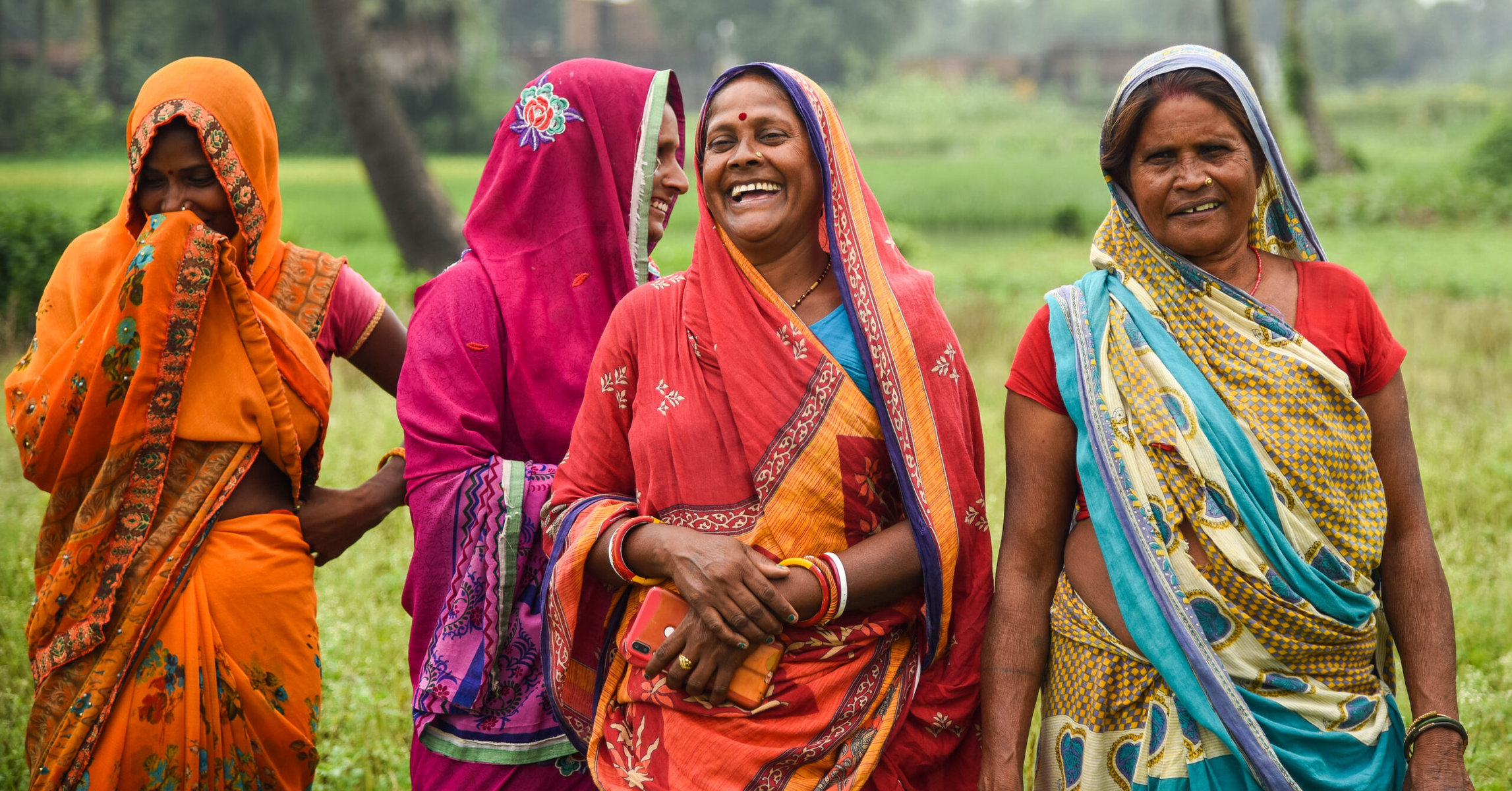 Women in Bihar are smiling in a field and wearing bright-colored clothing.