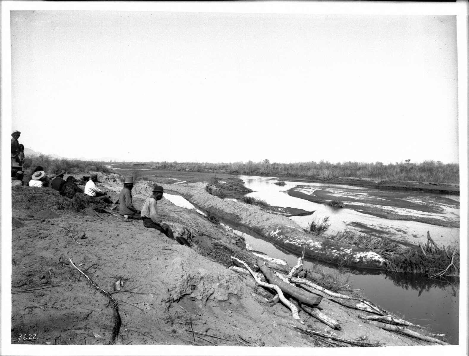 A black and white photo of a group of Pima farmers sit on the shore of the Gila River looking at an irrigation dam, circa 1900.