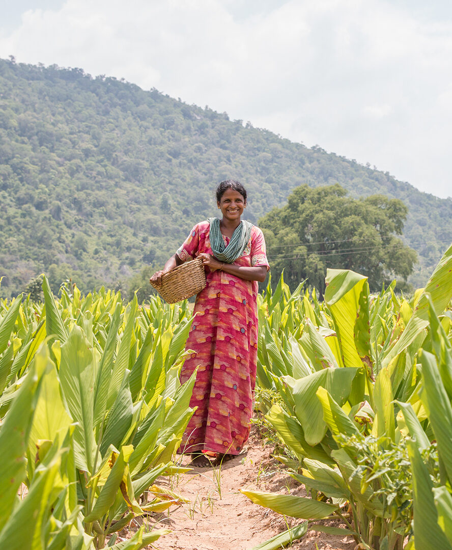 A farmer stands in a field in Tamil Nadu. She is smiling and holding a basket.