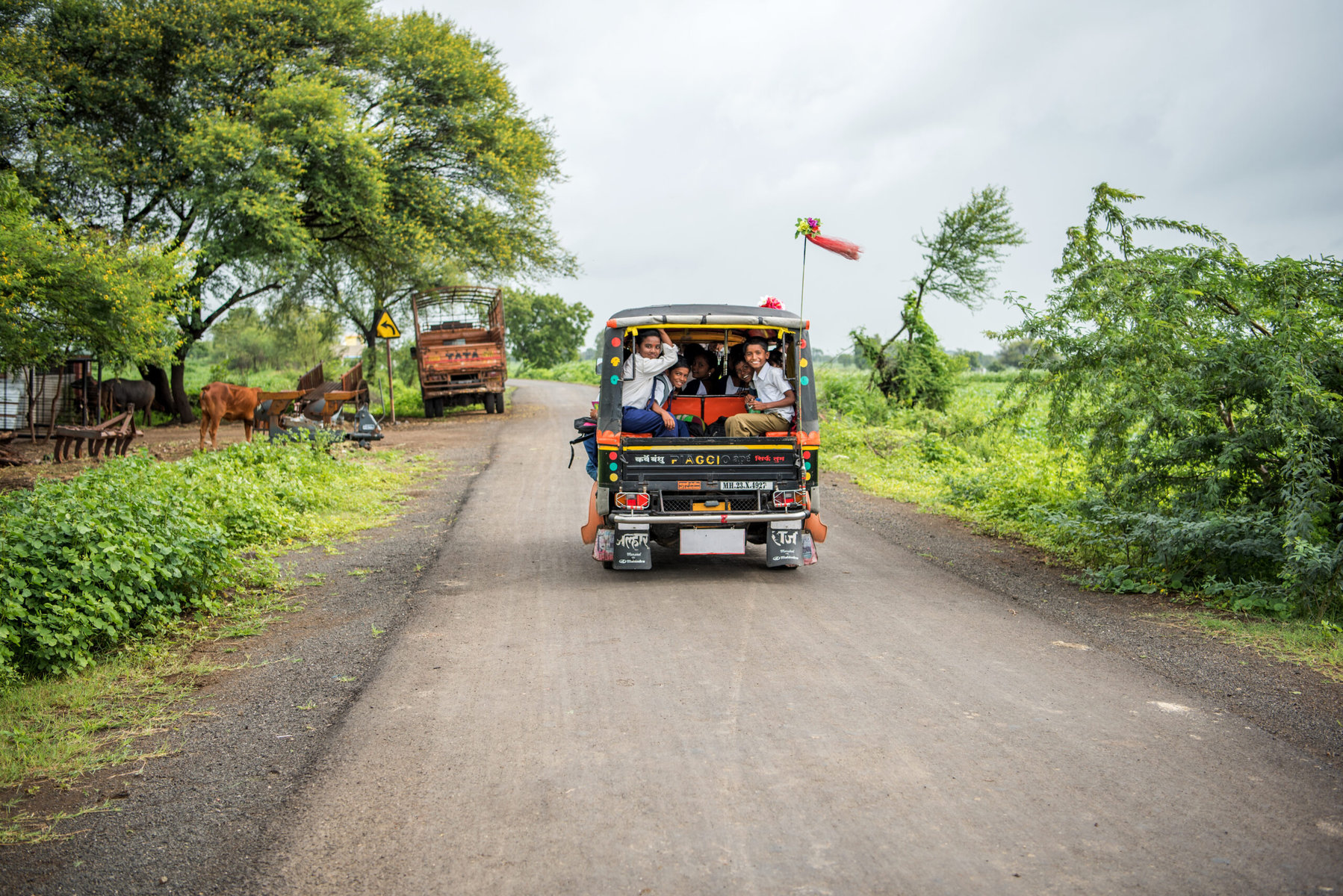Children on their way to school in a rural village in Beed. They are in an open-air vehicle on a dirt road with green all around.