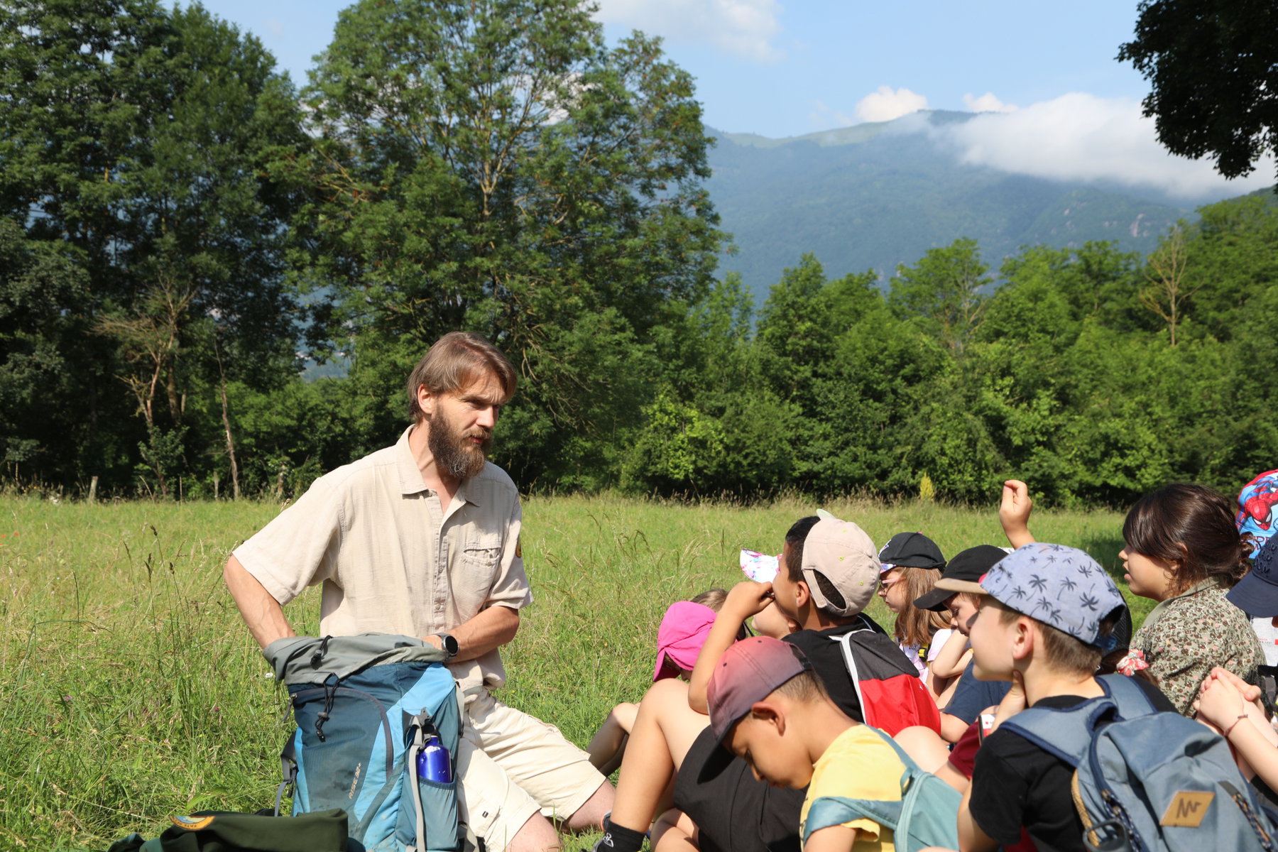 A mountain guide talks to a group of school children in a field with mountains in the background.
