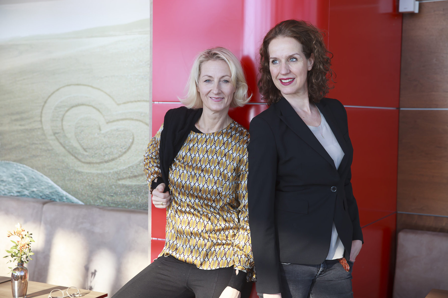 Angela Nelissen (left) and Christiane Haasis (right) pose in businesswear against a red background.