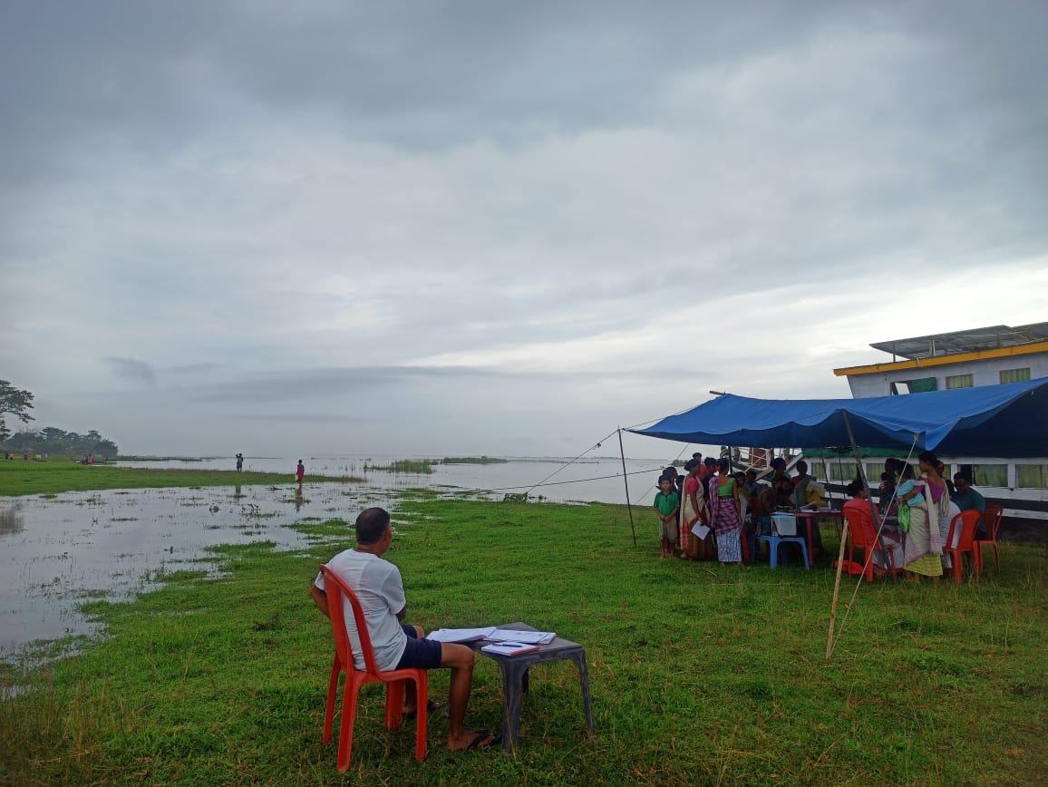 On a rainy day, a boat clinic conducts a camp in Majuli. A tent is set up in a marshy area by the water.