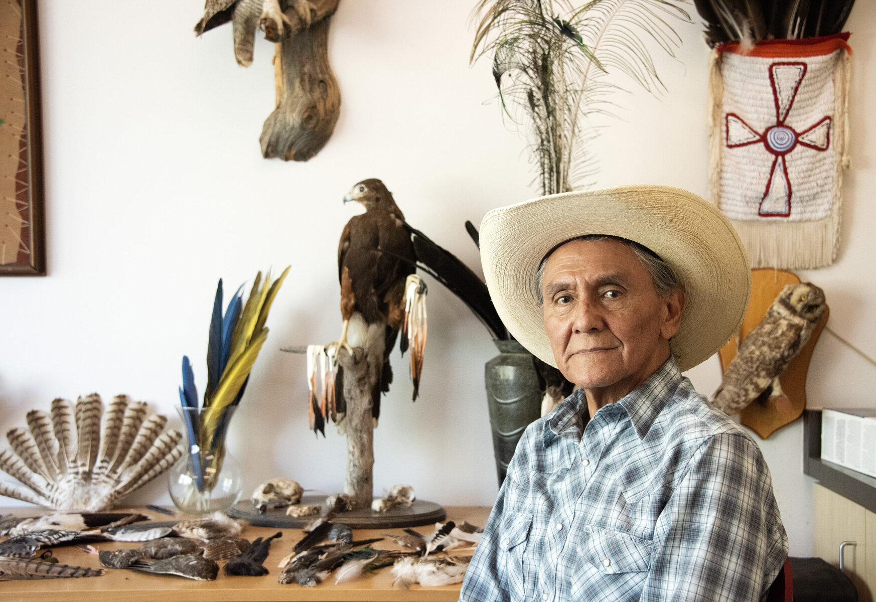 A man in a cowboy hat and plaid shirt sits in front of a table with feathers on it.