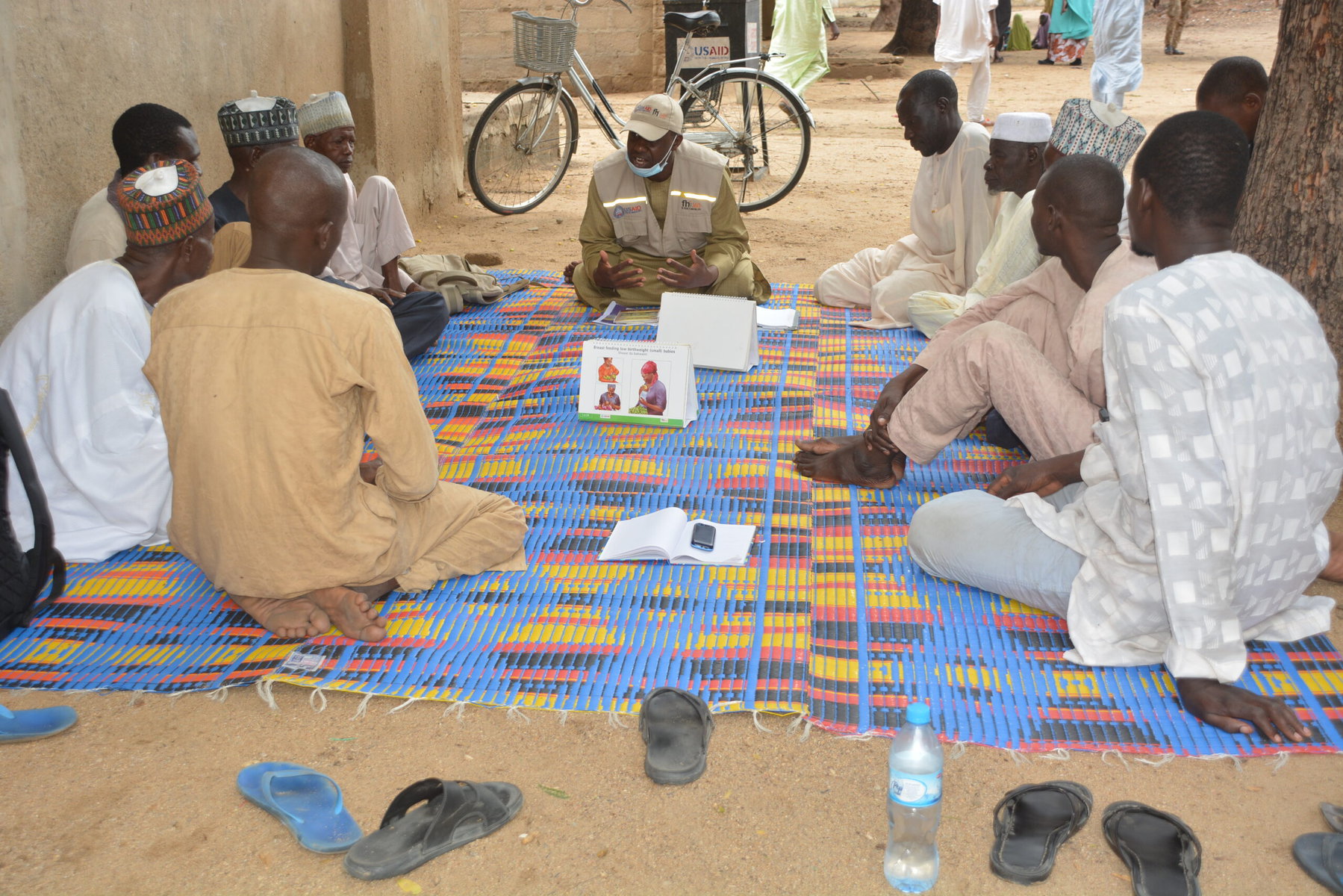 A group of fathers learns about maternal and child care. They sit together on a colorful rug.