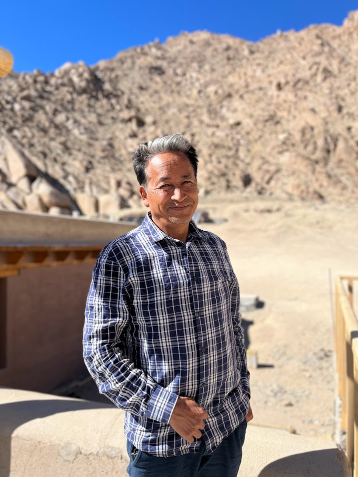 Sonam Wangchuk stands in a desert setting with a building behind him.