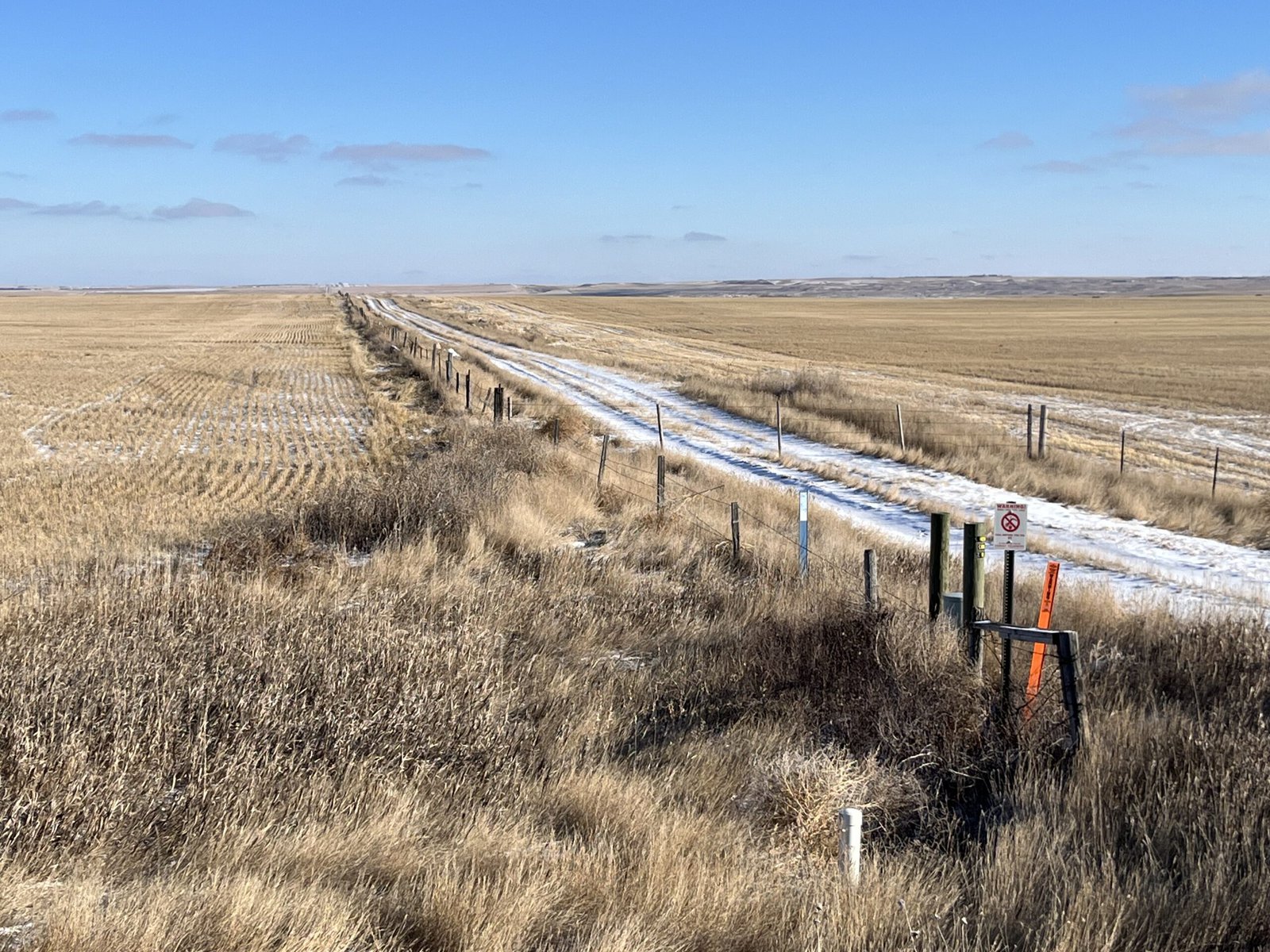 A snowy dirt road through fields, with a vertical white cylinder sticks up in the foreground. The vertical white cylinder is one of many that Amy Yoder with the Sheridan County Conservation District checks throughout the growing season.