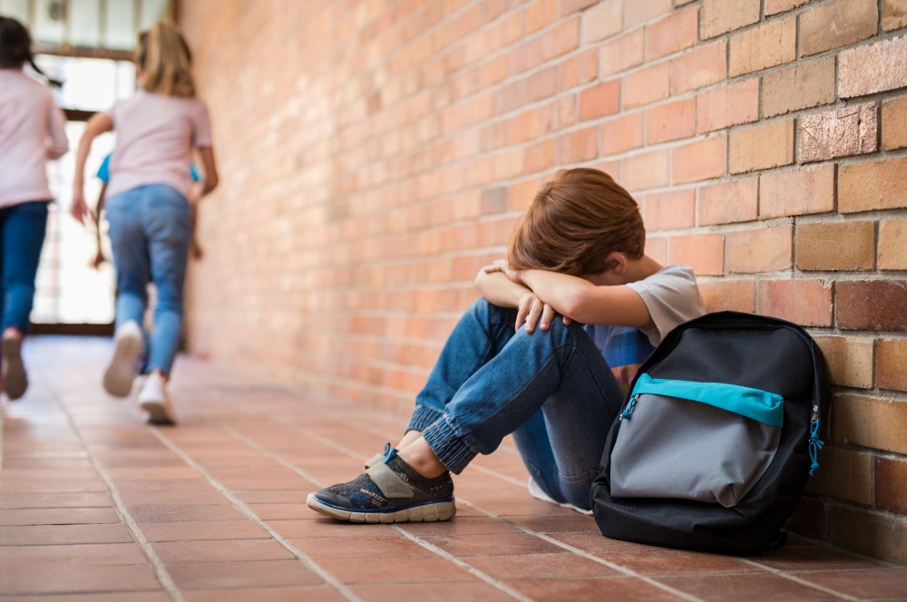 A boy sits alone with his head in his hands in a school hallway while other kids run away.