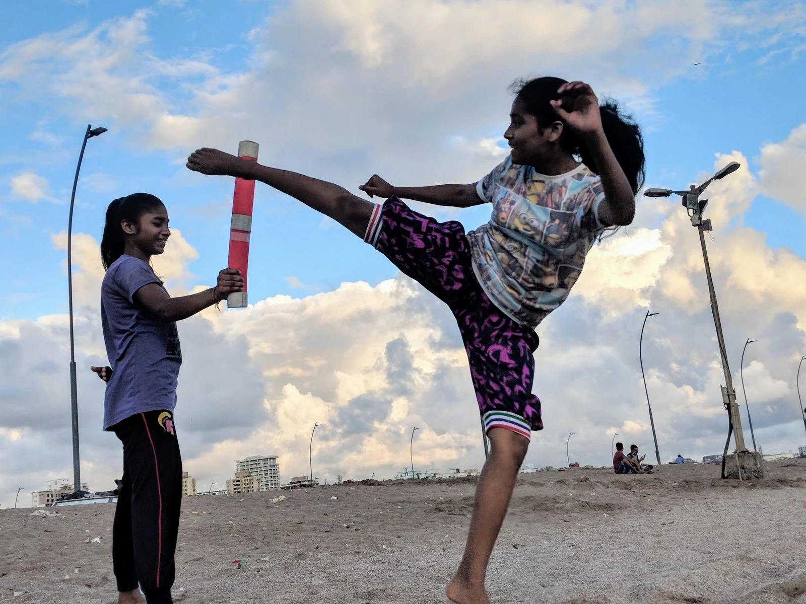 A girl kicks at an object held up by another girl on the beach.