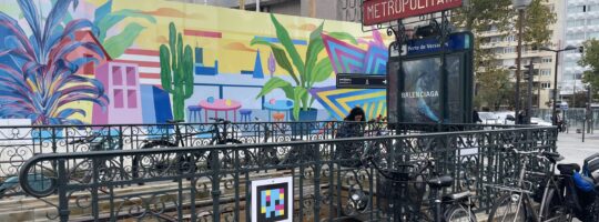 A Paris Metro entrance with a NaviLens code on the railing. There is a bright mural on the wall behind featuring plants.