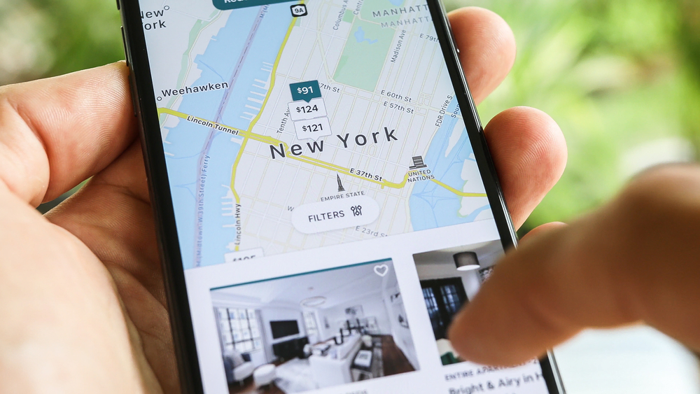 A hand holds a phone viewing New York Airbnb listings.