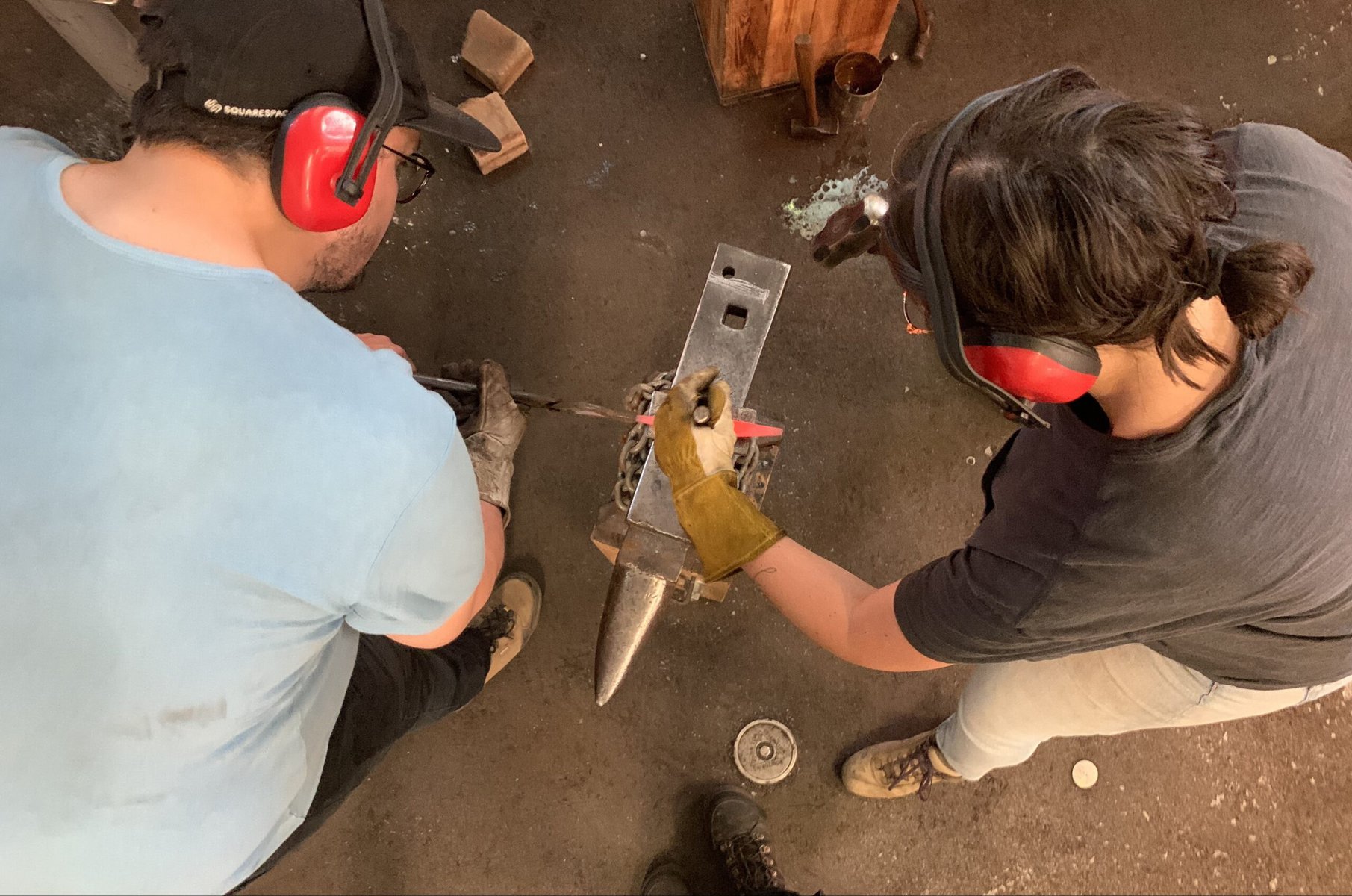 Two people wearing protective headphones and gloves work with metal, seen from above.