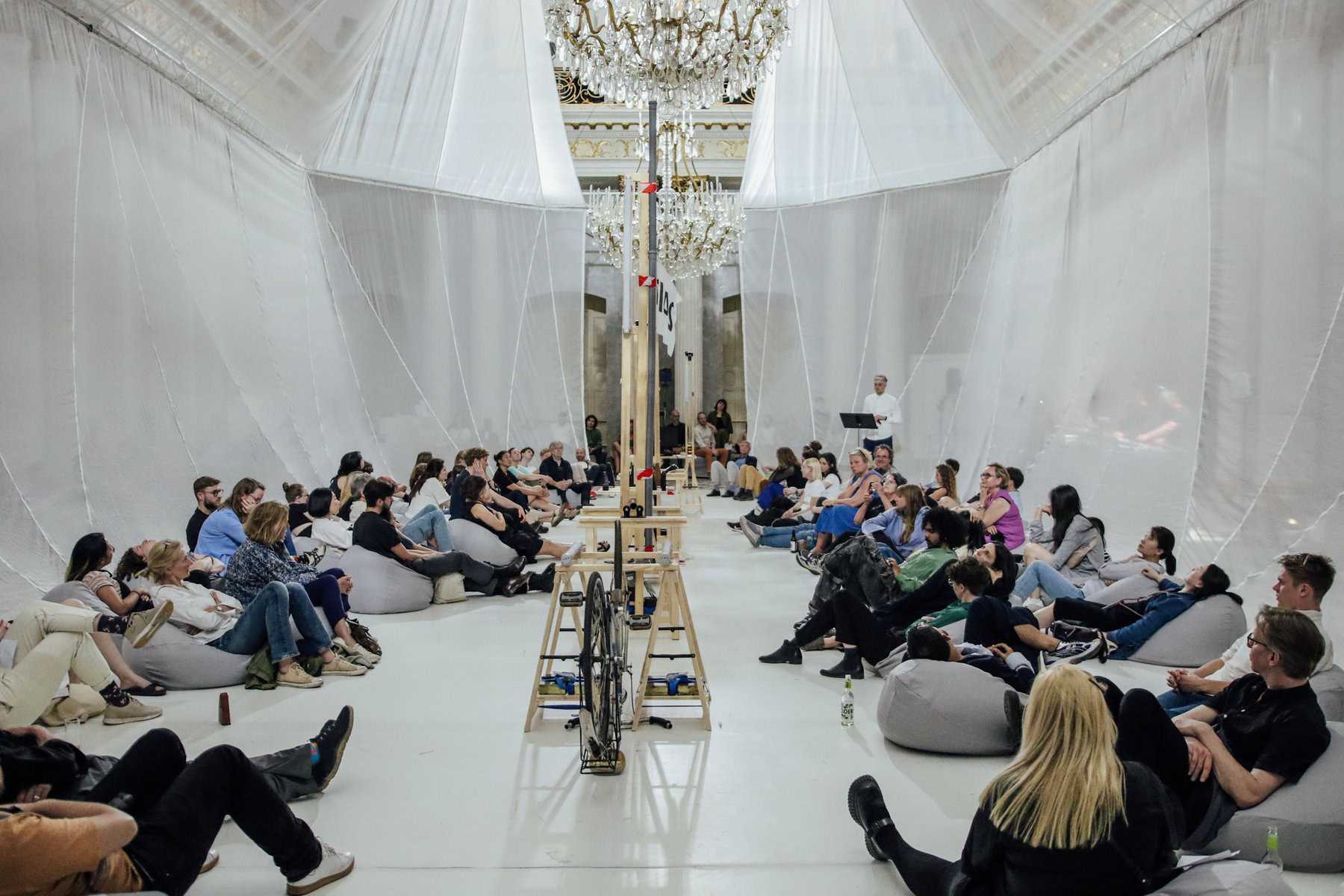 Listeners lounge on bean bags beneath chandeliers at a concert.