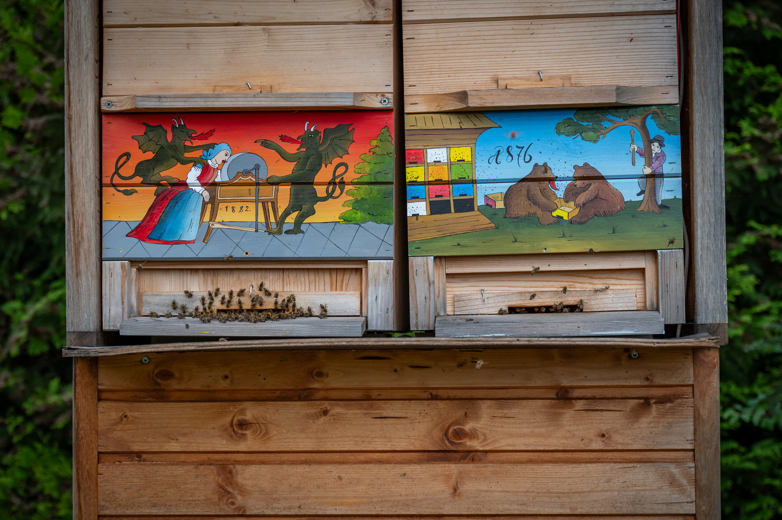 Wooden apiaries are painted in bright colors. One shows a woman with dragon-like creatures. One shows two bears outside an apiary.