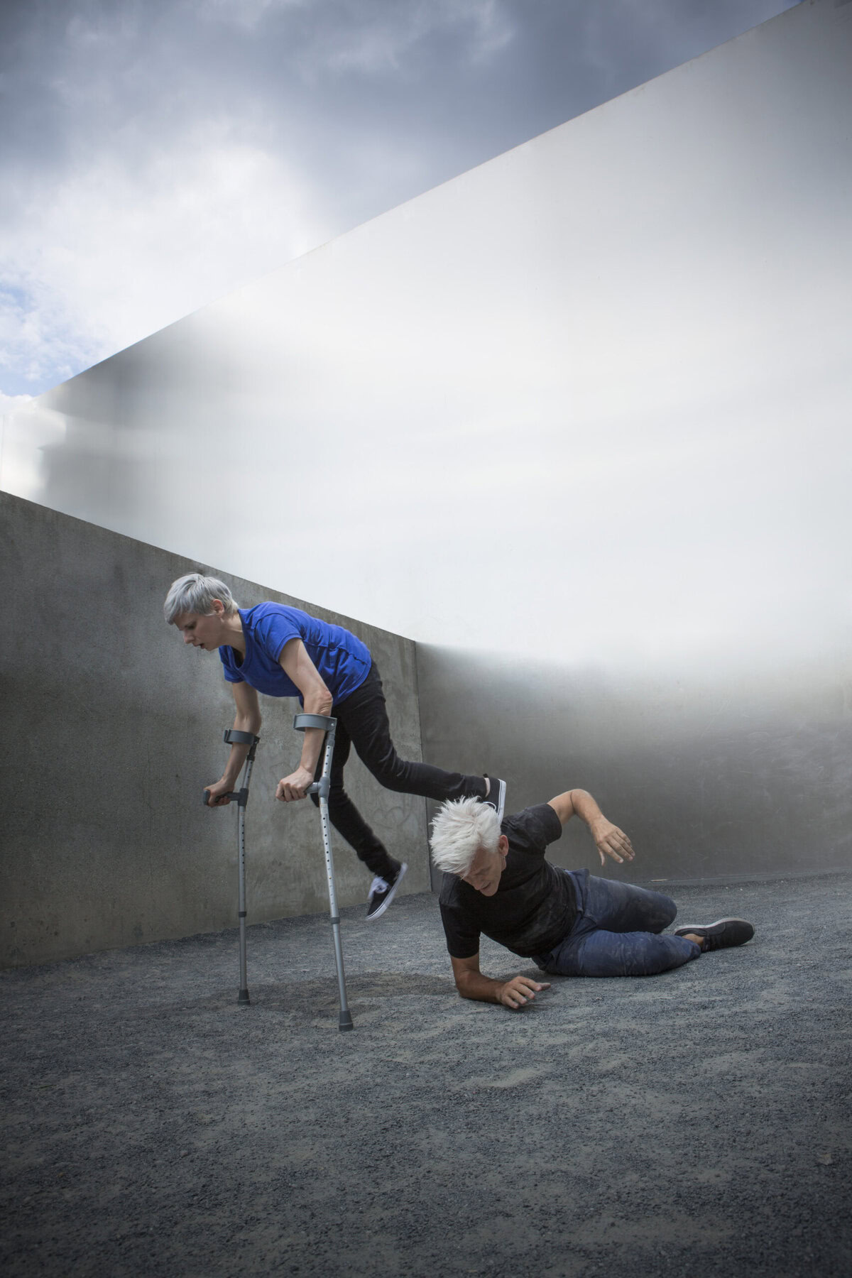 CUNNINGHAM (SLIGHT WITH SILVER HAIR AND BLUE TEE) BALANCES ON CRUTCHES WITH 1 FOOT ON NECK OF CURTIS, WHO LIES ON GRAVEL GROUND AGAINST CEMENT WALLS. (PHOTO: SVEN HAGOLANI)