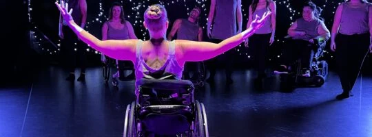 A woman in a wheel chair is facing the cast in on a dimly lit stage, her arms outstretched.