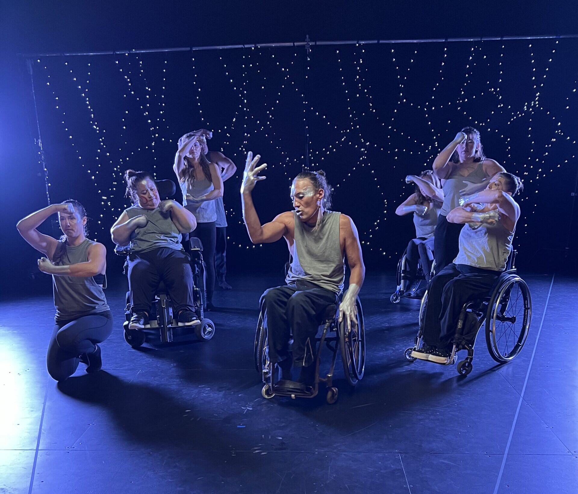 A group of dancers gesturing with their hands on a blue-lit stage. Some of the dancers are in wheel chairs and all have silver paint on their hands and faces.