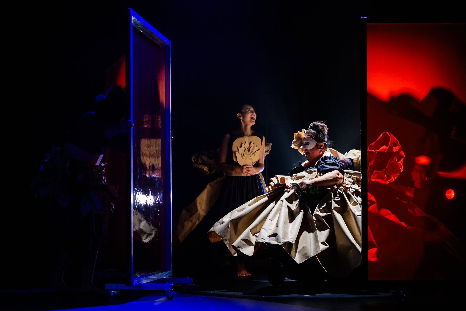 On a dramatically lit stage, a woman in wheelchair wearing an elaborate, wide-skirted dress and a mask looks into window. A woman behind her appears to laugh.