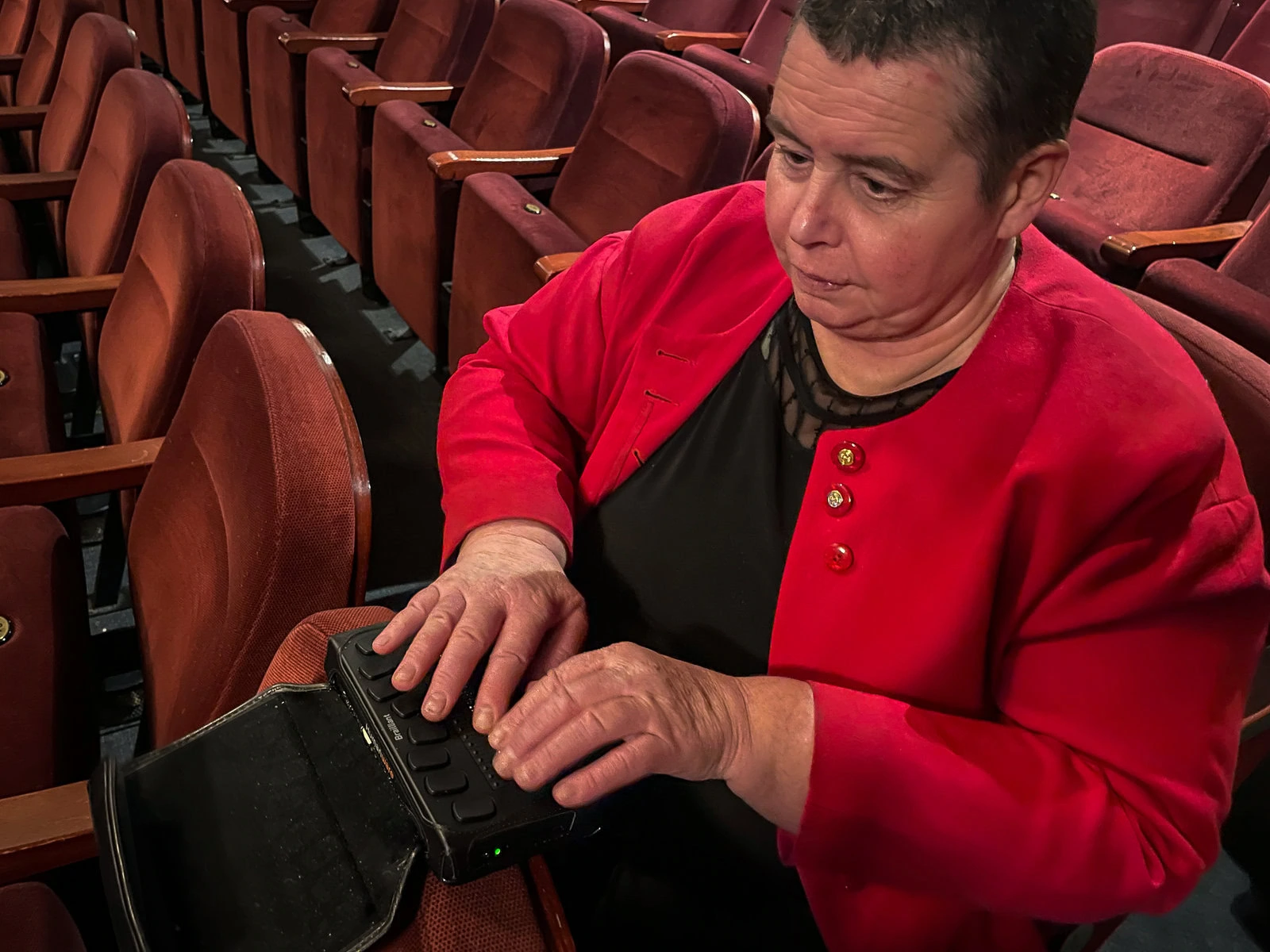 A woman in a red jacket sits in a row of audience seats with her hands on a Braille reader.