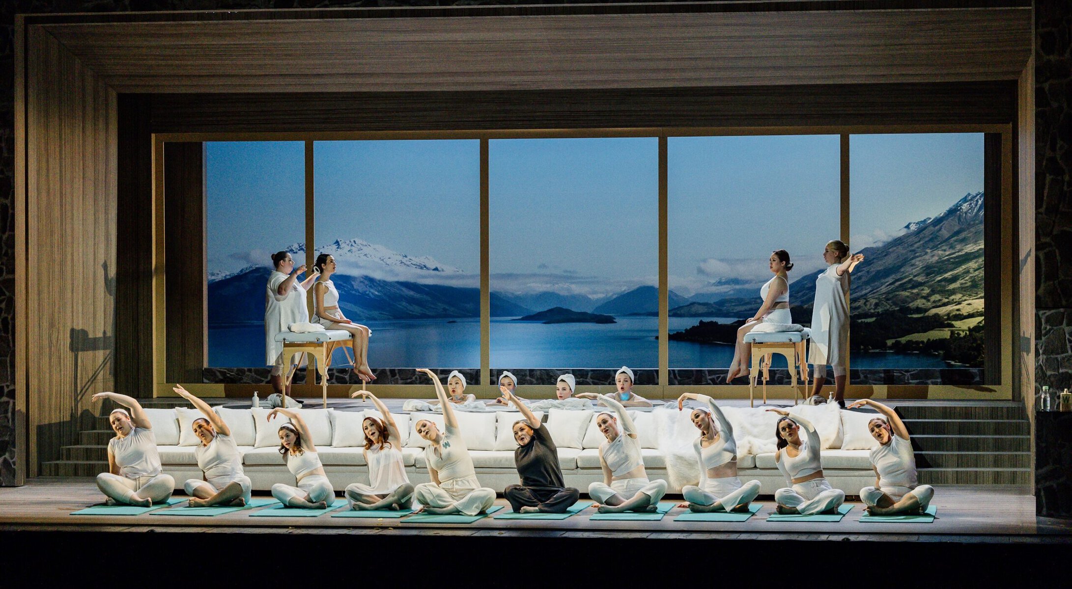 A large group of performers on stage in what appears to be a spa scene, some stretching on yoga mats, in front of a set of windows showing a mountain view.