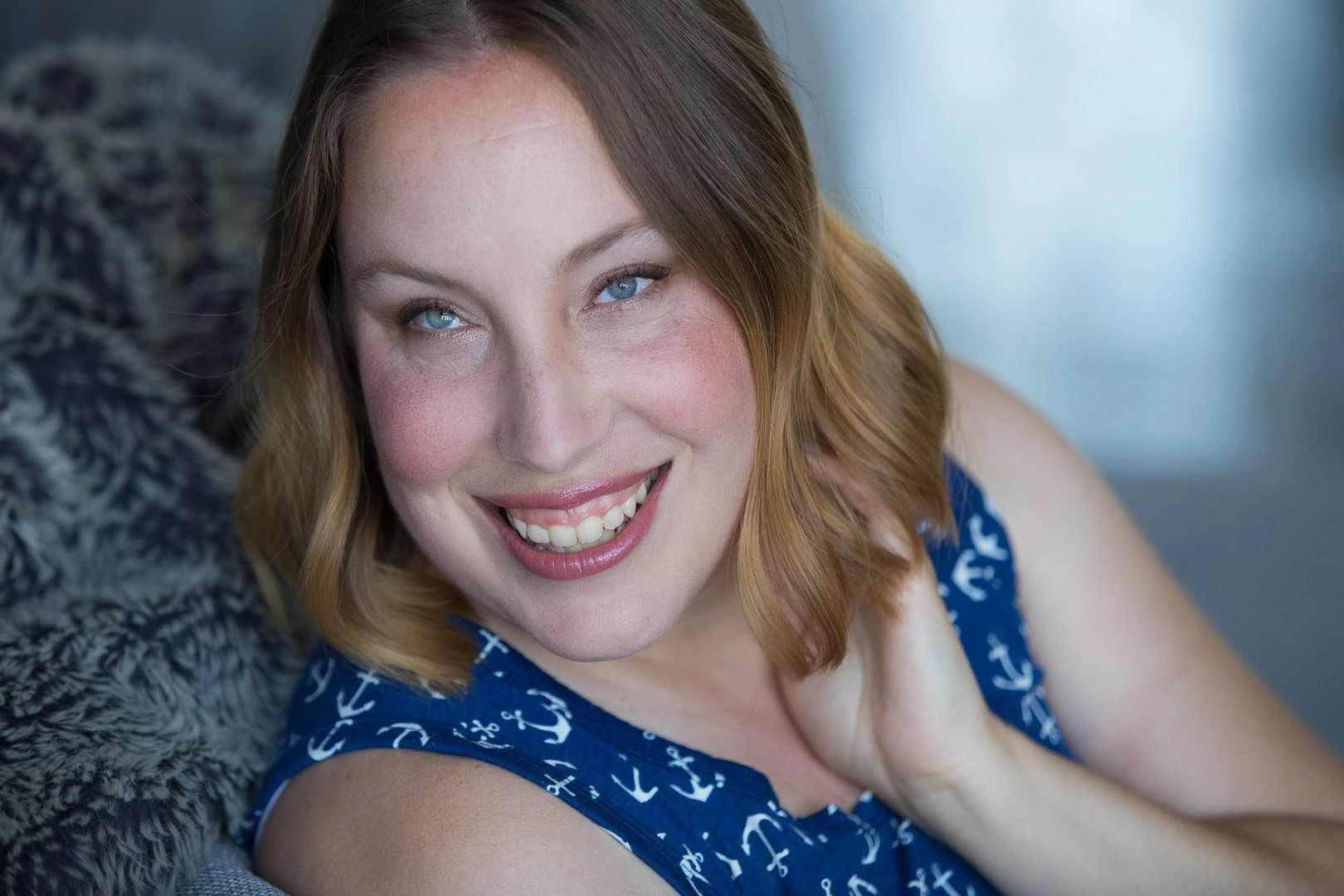 Headshot of a white woman in her 30s with green eyes, freckles and brown-blonde, curly hair, Smiling broadly with her hand curled up next to her face, wearing a sleeveless anchor print top. Photo credit Cheshire Isaacs of Oaktown Boudoir.