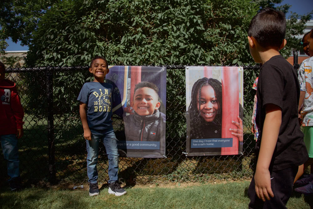 A young student poses in front of his photo hanging from a fence.