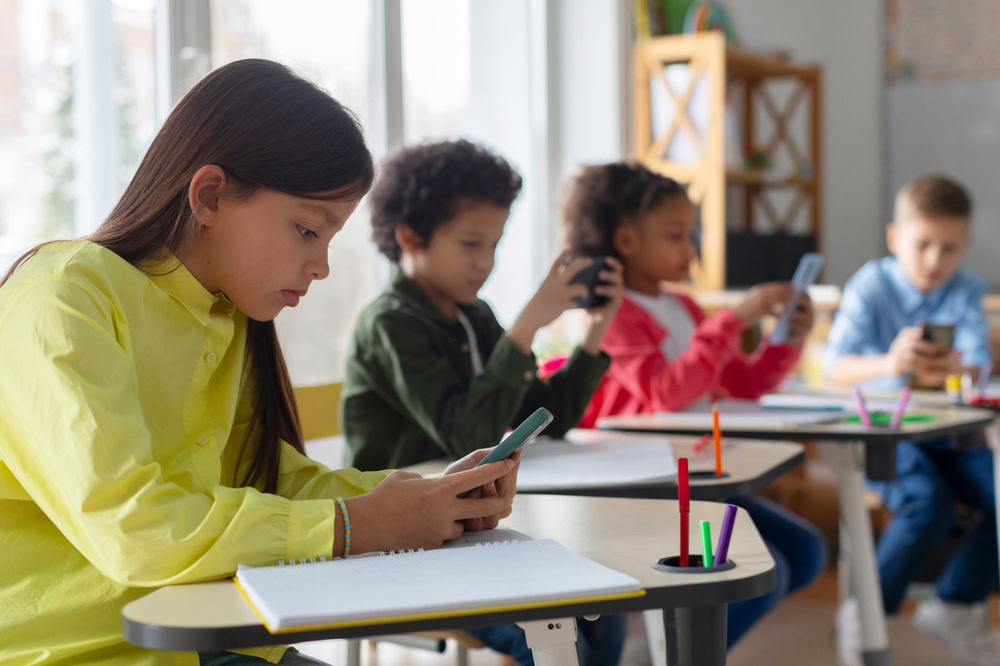 Kids using smartphones, sitting at desks in a classroom.