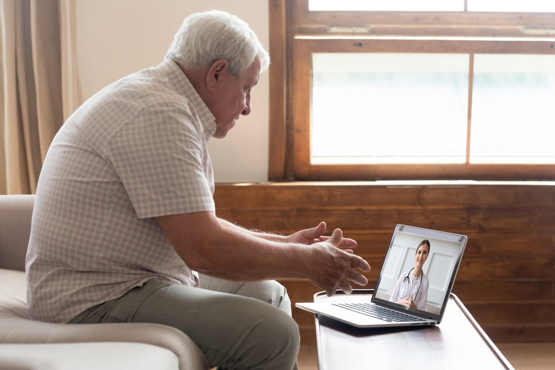 Elderly man seated on a sofa in a telehealth appointment on his laptop.