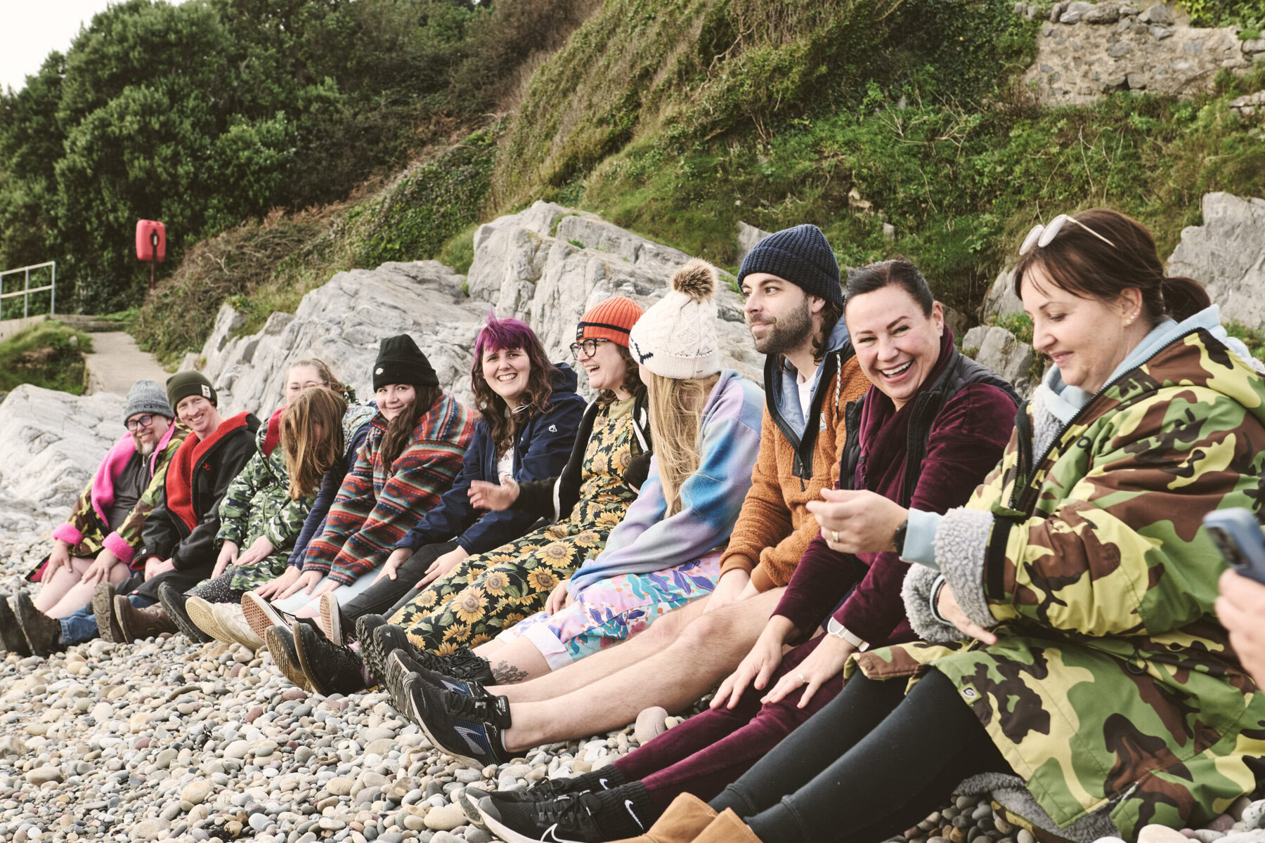 A group of swimmers, fully clothed, sitting on a rocky beach.