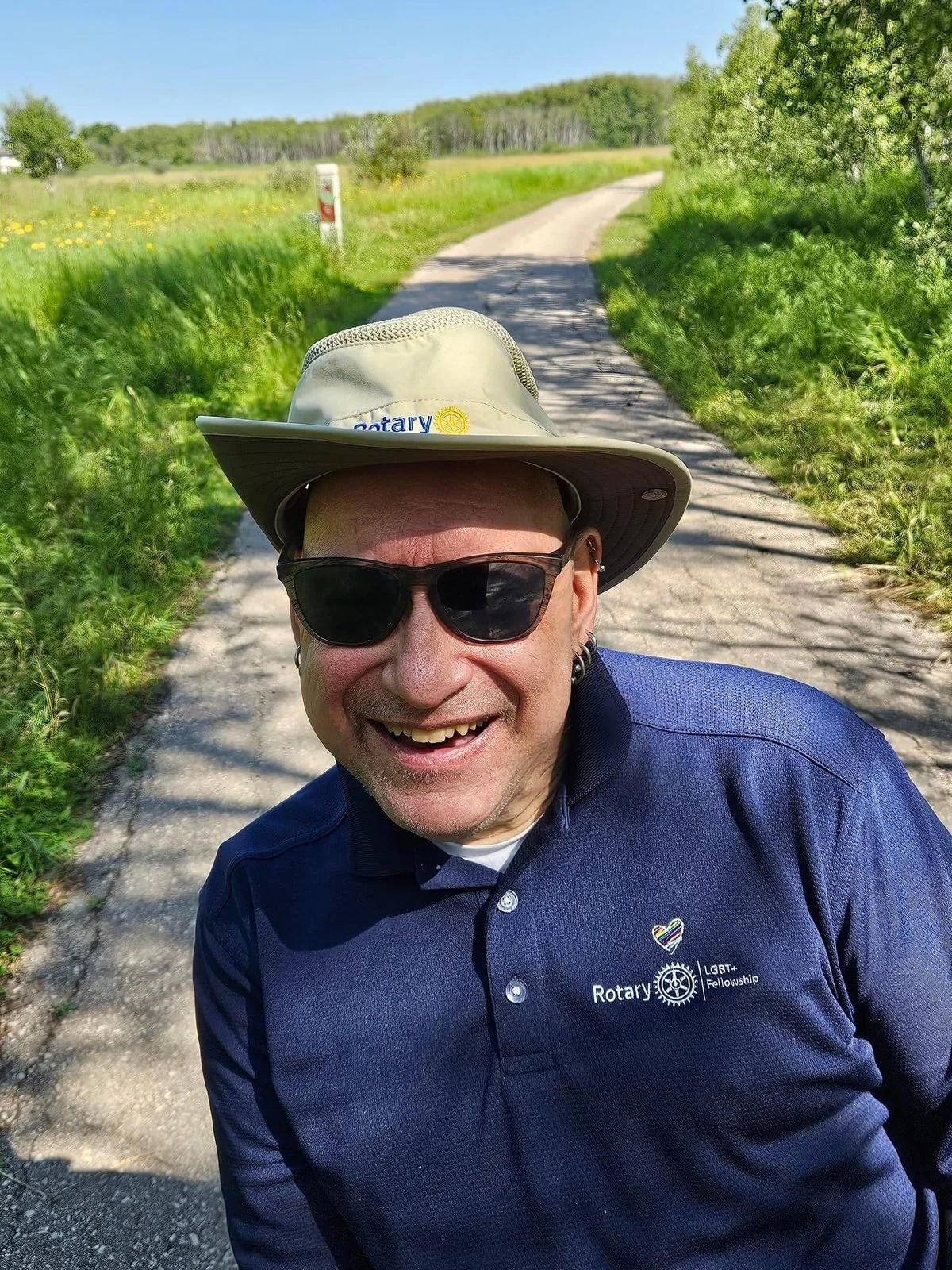 Peter Tonge poses while exploring Winnipeg’s Assiniboine Forest. He wears a hat and sunglasses and an accessible trail surrounded by grass is visible behind him.