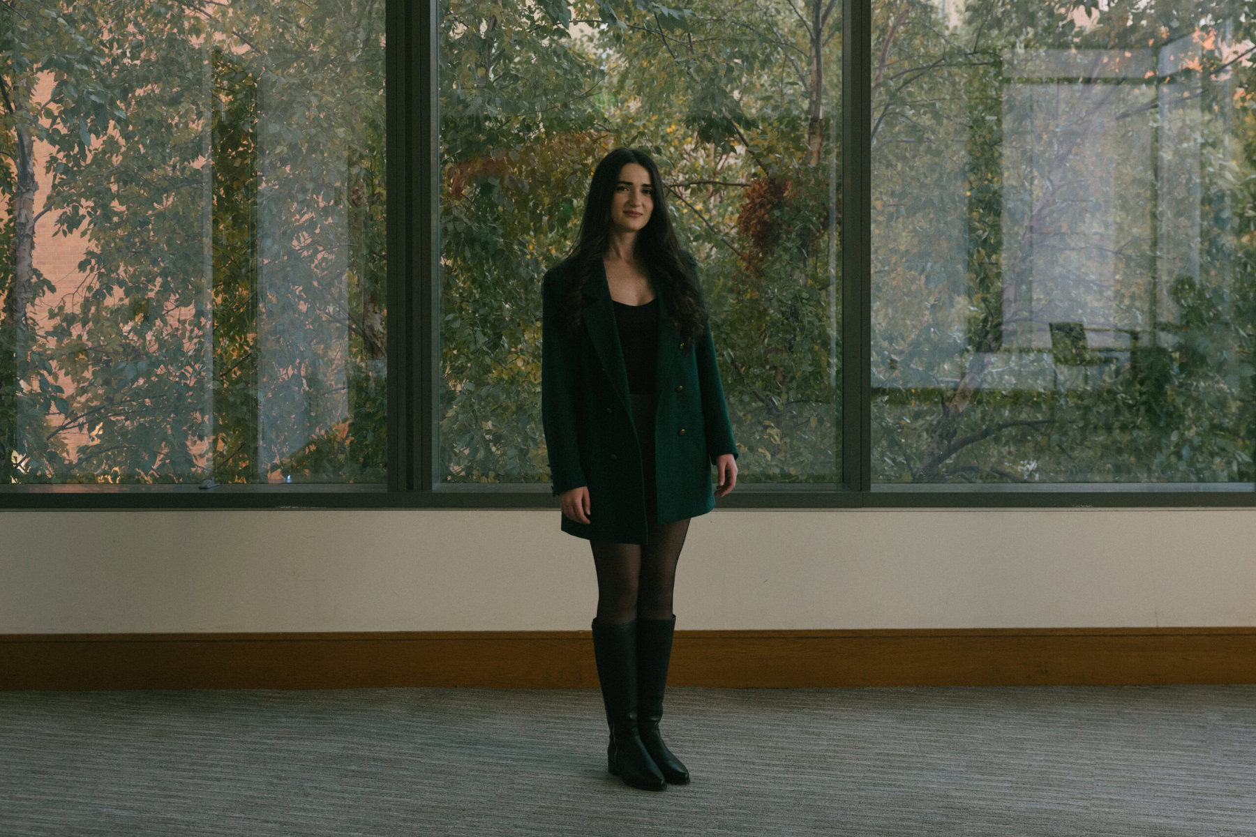 Tamari Natelauri poses for a portrait on campus at Drexel University, in Philadelphia. She is wearing a skirt and boots and a green jacket and standing in front of a window with trees behind them.