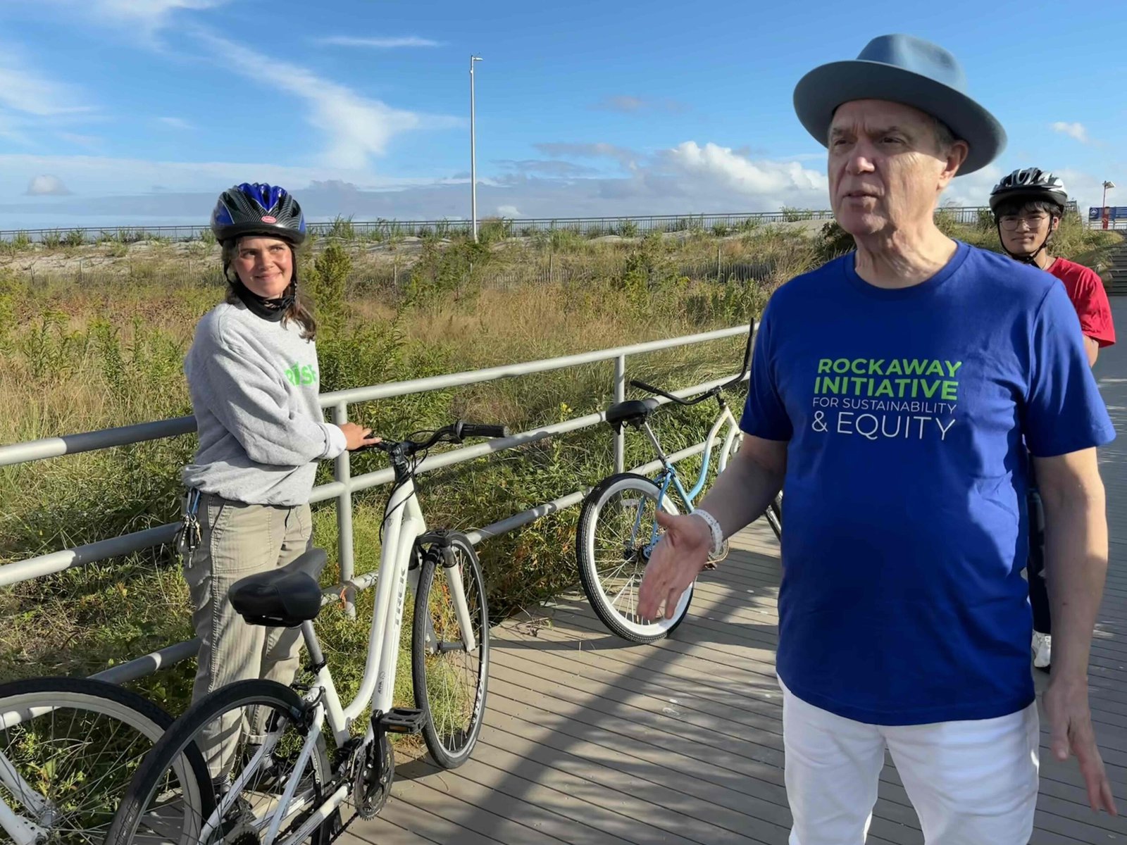 David Byrne stands on a boardwalk in Rockaway with a young person standing near a bike.