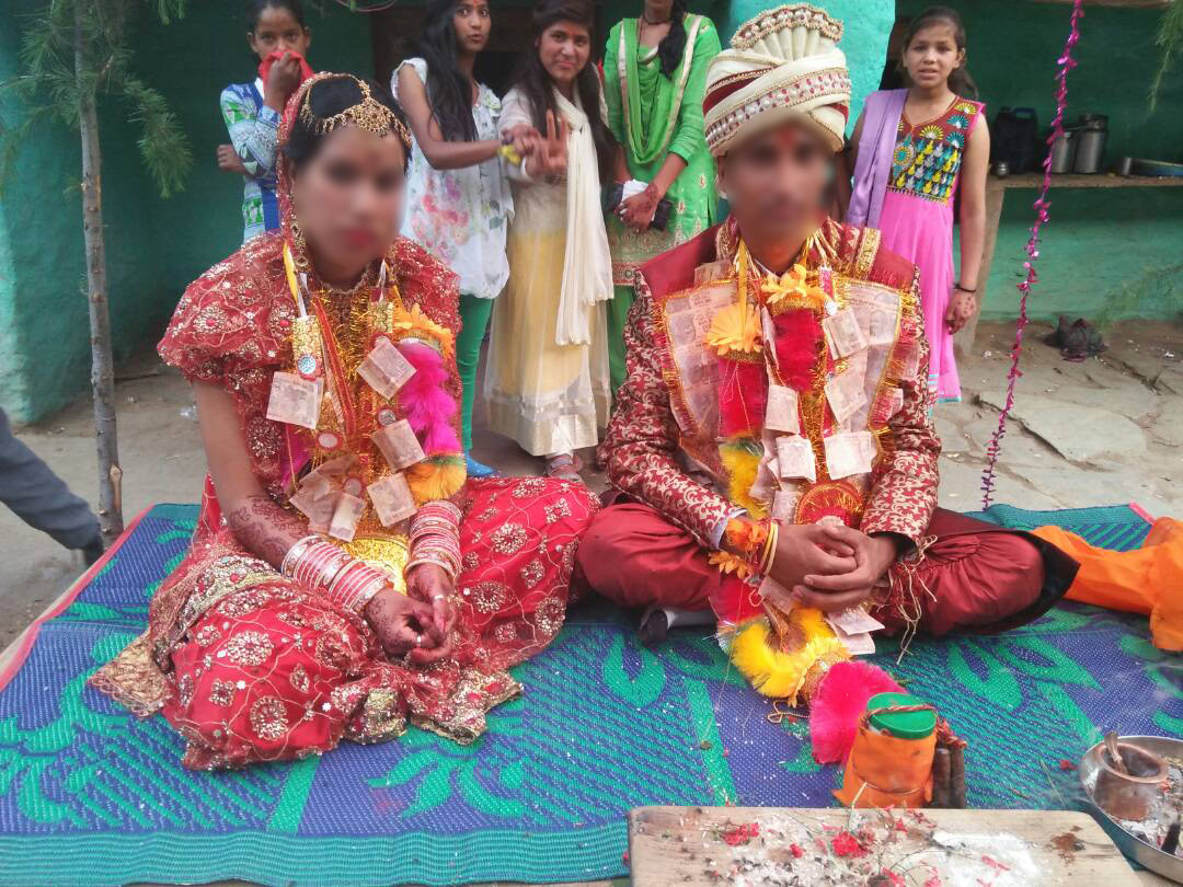 A bridge and groom are seated on a mat wearing bright red outfits.