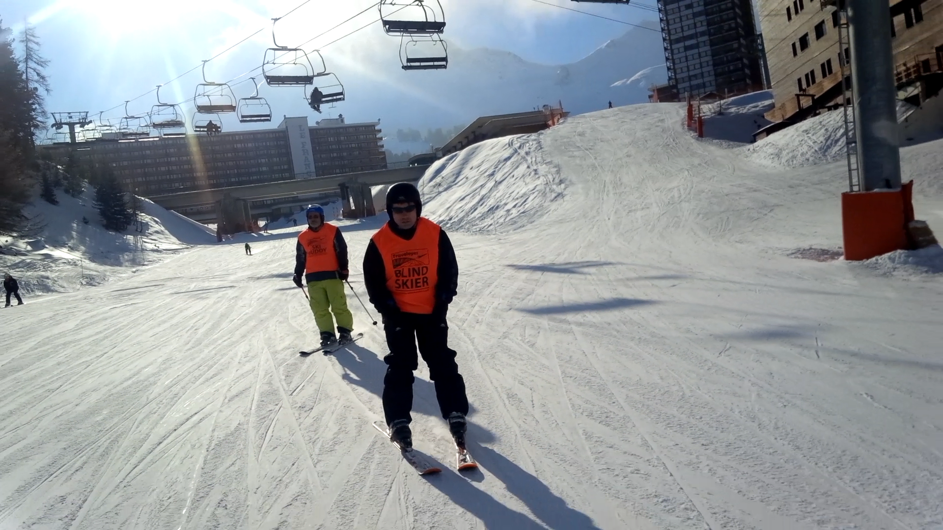 Two skiers wearing bright orange vests on a ski trail with a chairlift above them.