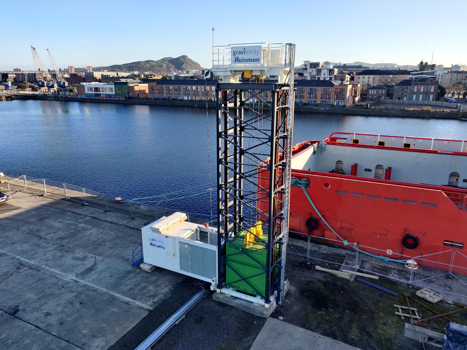 The Gravitricity demonstrator with a waterway and buildings behind it. It looks like a scaffolding tower with a white platform on top.