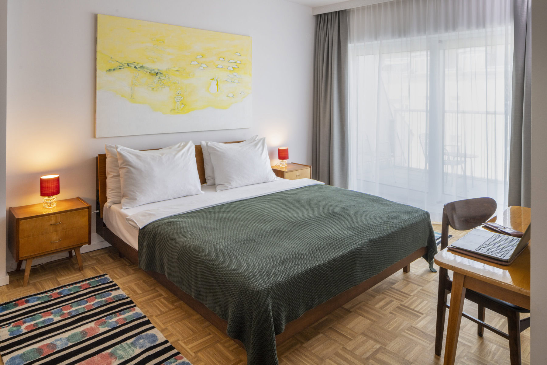 A bedroom with light coming through the window. There is a green bedspread and a yellow painting behind the bed.