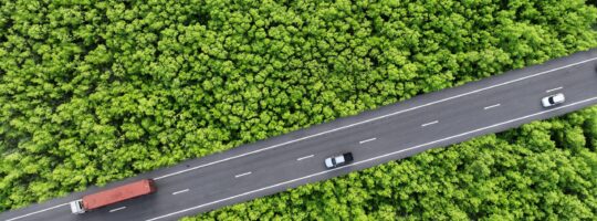 An aerial view of a truck and cars driving on a straight road with forest on both sides.