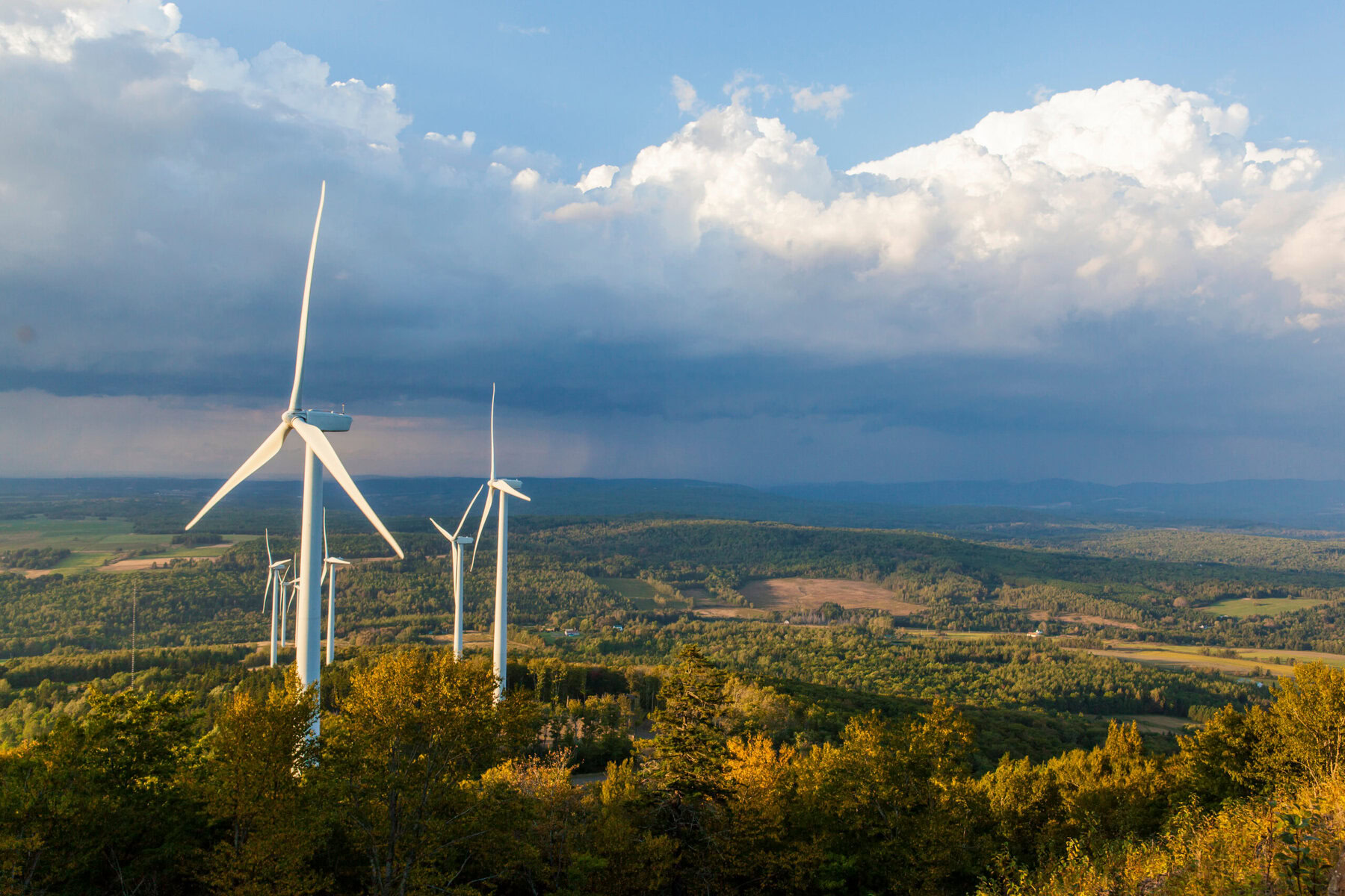 Wind turbines on a hill in Maine with a thunderstorm visible in the distance.