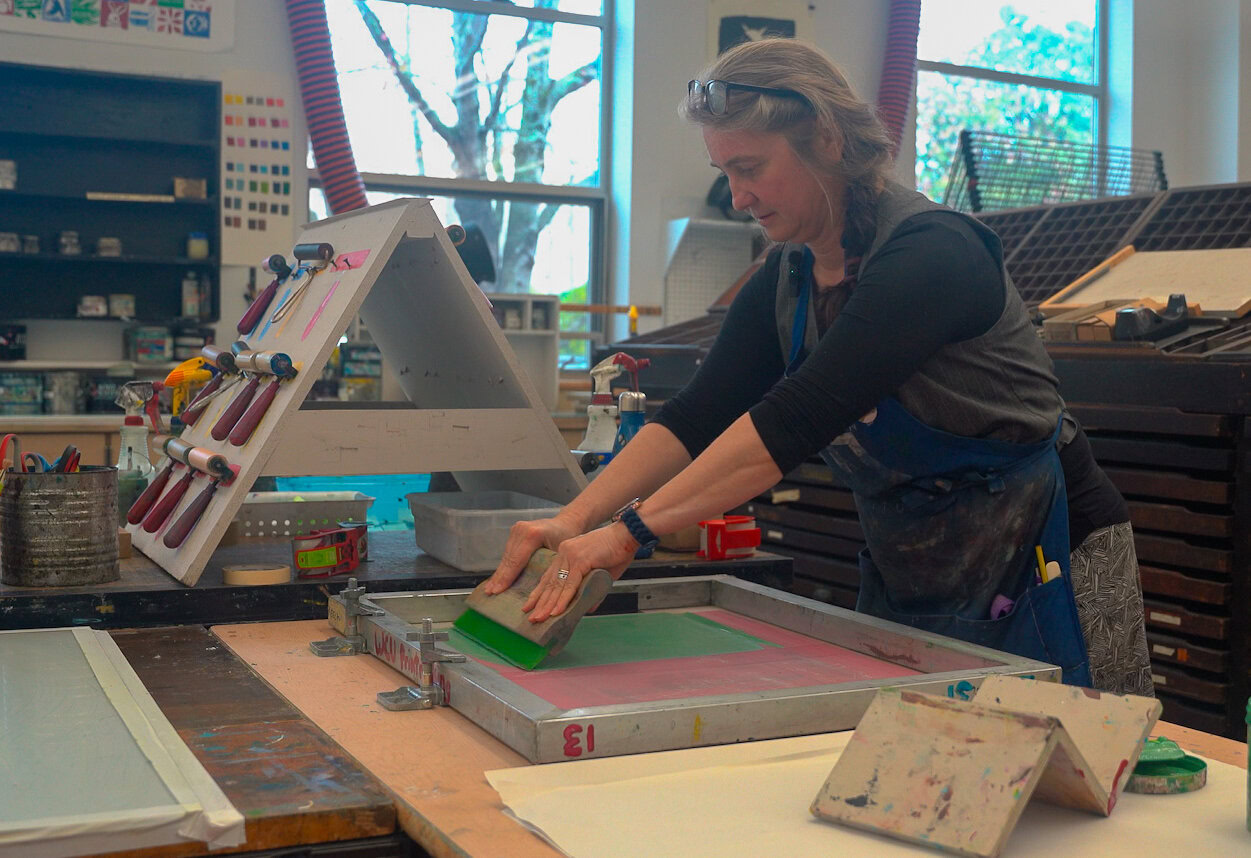 Tatiana Potts doing screen printing at a table with tools. She wears a green apron.