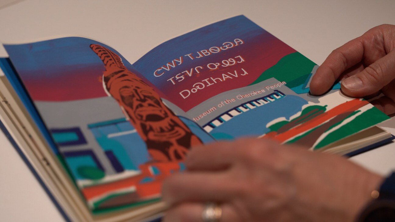 Hands hold a book open to a page with a colorful illustration and writing in both English and Cherokee that reads "Museum of the Cherokee People."