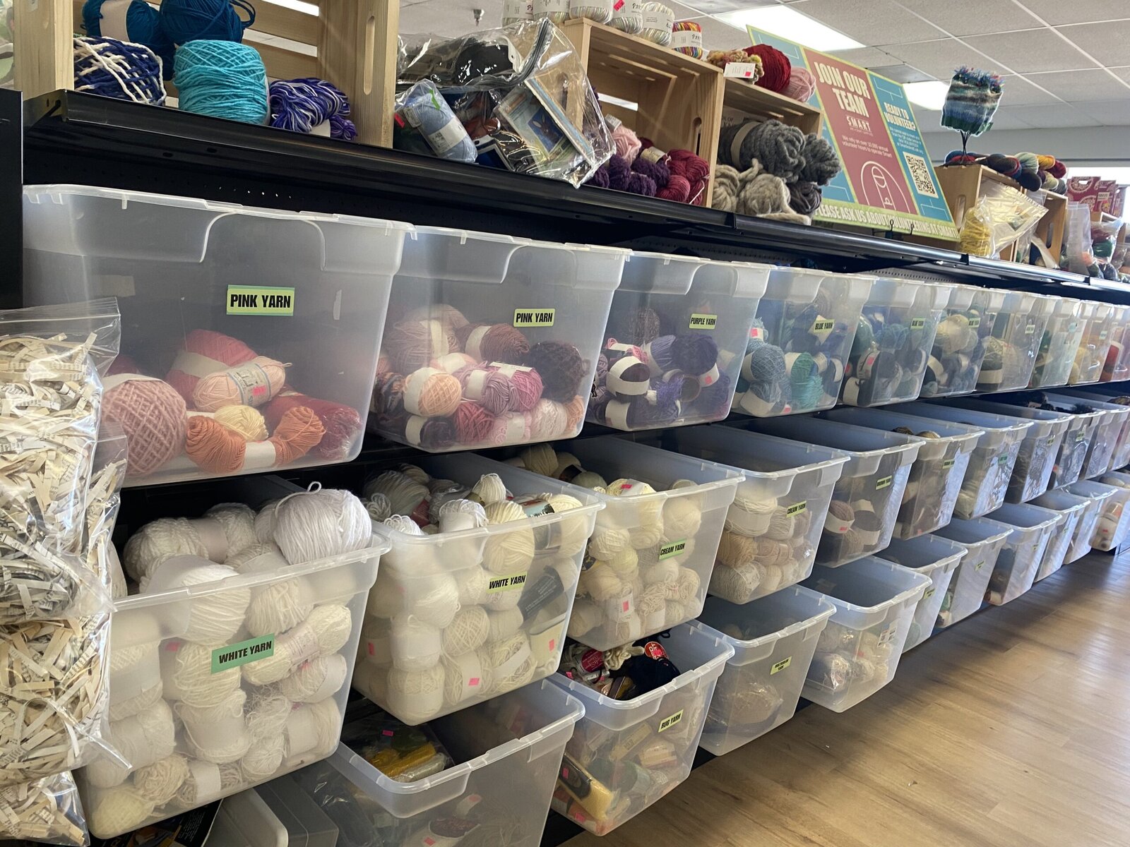 Plastic containers of yarn in all different colors are in orderly rows on three shelves.