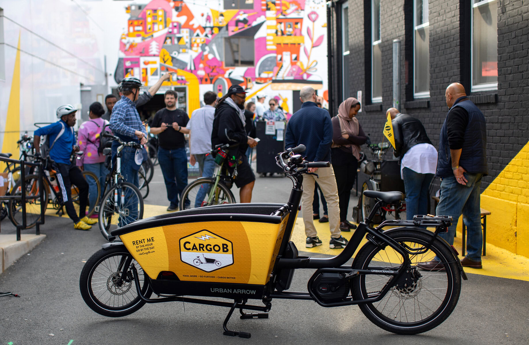 In a paved outdoor area between buildings, a CargoB bike is in the foreground, groups of people talk in the background with a colorful mural behind them.