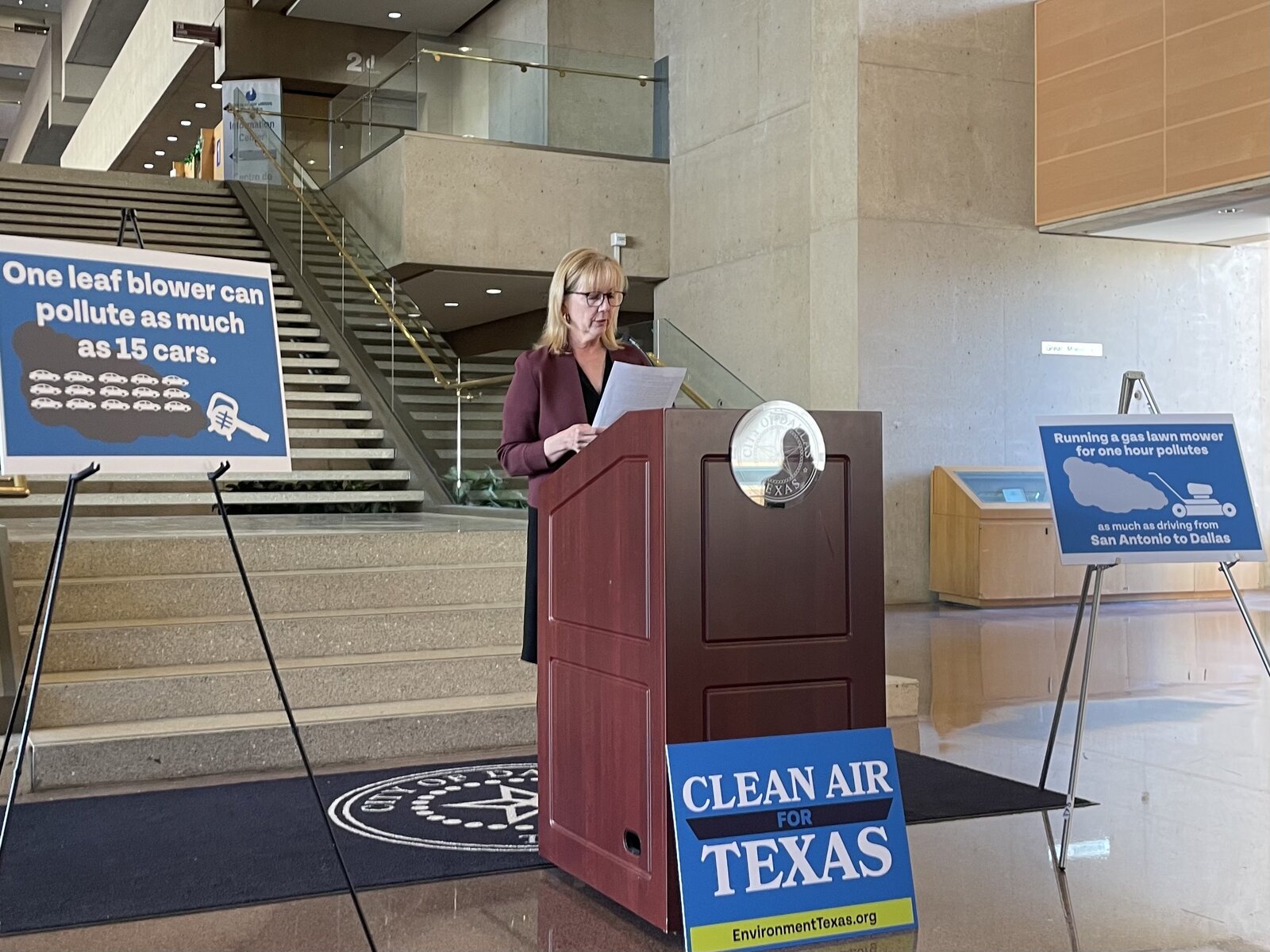 Dallas Councilwoman Kathy Stewart, chair of the council’s Parks, Trails, and the Environment committee, speaks about electric lawn care at event about North Texas air quality. She stands behind a podium in a government building. Signs around her promote clean air and electric lawn equipment.
