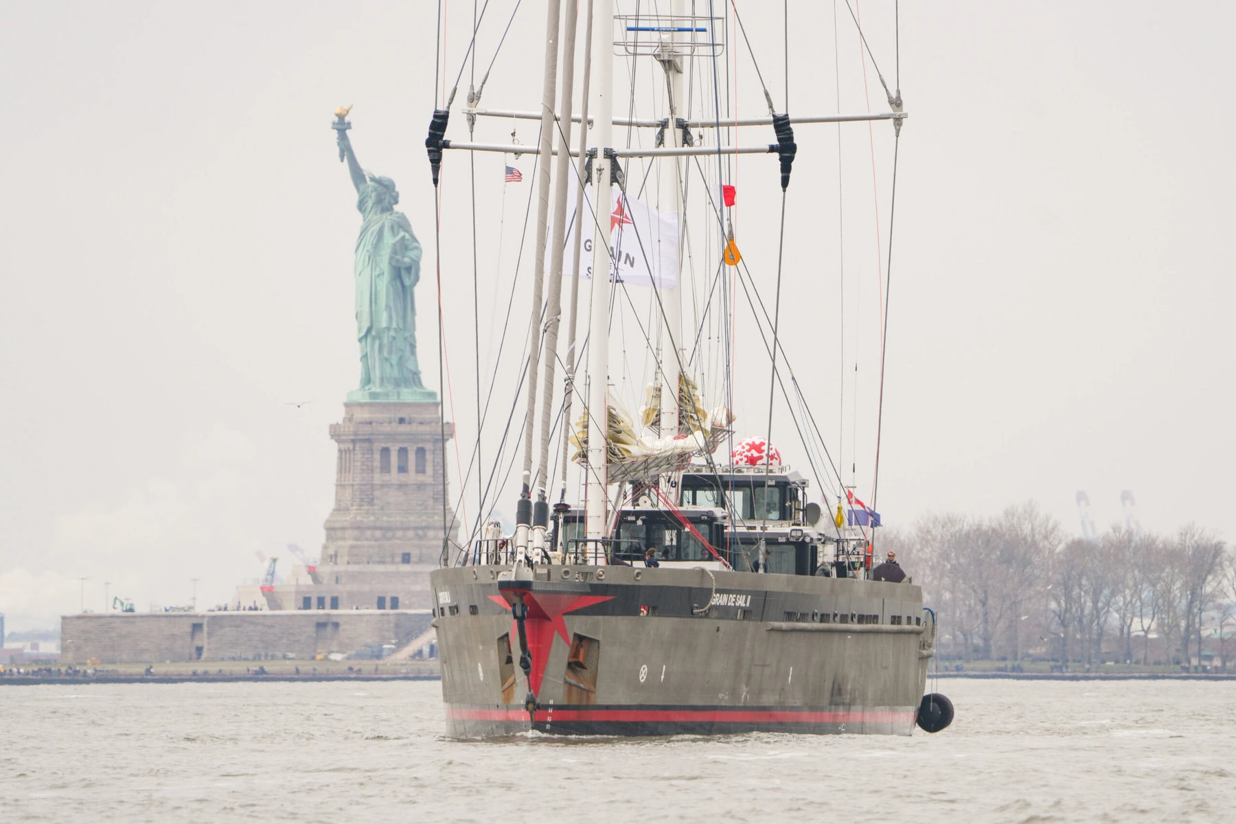 A sailing ship passes in front of the Statue of Liberty on a cloudy day.