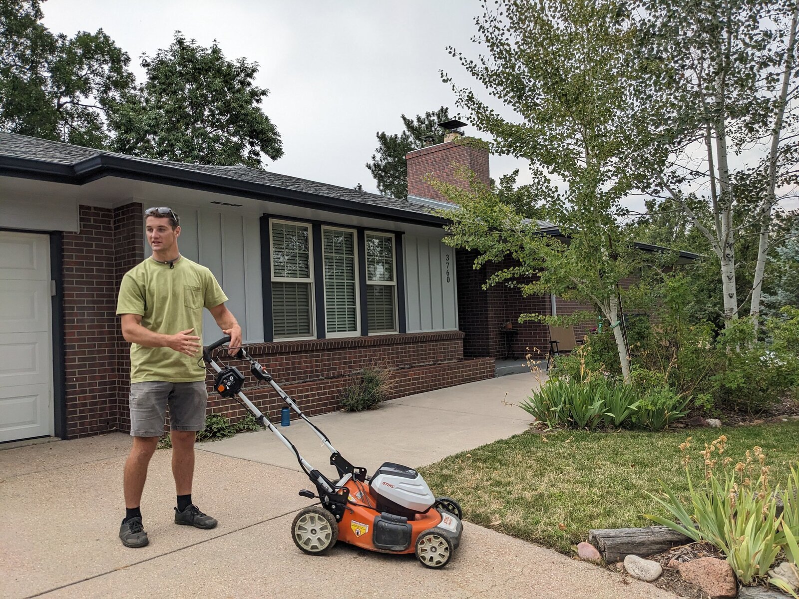 A man gestures while standing behind an electric lawn mower in a driveway in front of brick home.