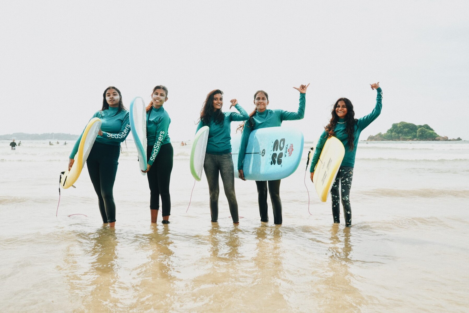 A group of young women pose on the beach with surf boards.