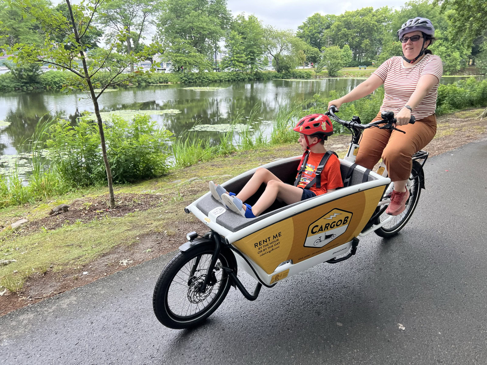 A mother rides a cargo e-bike in a park while a child rides in the cargo compartment. There is a pond in the background.